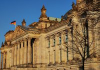 Das Bild zeigt die Fassade des Reichstagsgebäude in Berlin. Im Hintergrund ist ein blauer Himmel zu sehen. Auf dem Dach weht die deutsche Flagge. Die Architektur ist reich verziert mit Säulen und Skulpturen, die die historische Bedeutung des Gebäudes unterstreichen.