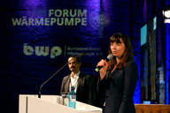 Eine Frau spricht am Mikrofon auf einer Bühne, während ein Mann im Hintergrund steht. Im Hintergrund sind Logos des Forums Wärmepumpe und des Bundesverbands Wärmepumpe sichtbar. Die Szene hat eine professionelle Atmosphäre mit blauer Beleuchtung.