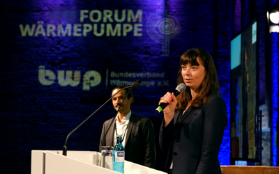 Eine Frau spricht am Mikrofon auf einer Bühne, während ein Mann im Hintergrund steht. Im Hintergrund sind Logos des Forums Wärmepumpe und des Bundesverbands Wärmepumpe sichtbar. Die Szene hat eine professionelle Atmosphäre mit blauer Beleuchtung.