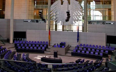Ein Blick auf den Plenarsaal des Deutschen Bundestages. Stühle in lila sind im Halbkreis angeordnet, während im Hintergrund ein großer Bundesadler und die Flaggen Deutschlands und der Europäischen Union zu sehen sind. Der Raum ist modern und lichtdurchflutet.
