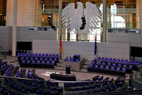 Ein Blick auf den Plenarsaal des Deutschen Bundestages. Stühle in lila sind im Halbkreis angeordnet, während im Hintergrund ein großer Bundesadler und die Flaggen Deutschlands und der Europäischen Union zu sehen sind. Der Raum ist modern und lichtdurchflutet.