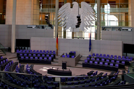 Ein Blick auf den Plenarsaal des Deutschen Bundestages. Stühle in lila sind im Halbkreis angeordnet, während im Hintergrund ein großer Bundesadler und die Flaggen Deutschlands und der Europäischen Union zu sehen sind. Der Raum ist modern und lichtdurchflutet.