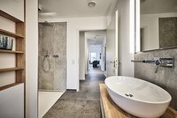 Ein modernes Badezimmer mit einer Dusche auf der linken Seite und einem eleganten, ovalen Waschbecken auf einem Holzregal. Im Hintergrund ist ein Flur zu sehen, der zu einem Wohnbereich führt. Helle Wände und großzügige Fenster sorgen für eine einladende Atmosphäre.