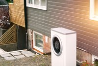 Eine weiße Klimaanlage steht neben einem Haus mit grauer Außenwand. Im Hintergrund sind Fenster sichtbar. Der Boden besteht aus Steinen und Erde. Pläne und ein Abflussrohr sind am Gebäude zu sehen. Es ist ein heller, sonniger Tag.