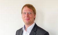 Ein junger Mann mit blonden Haaren trägt eine Brille und einen Anzug. Er lächelt freundlich und steht vor einer hellen, einfarbigen Wand. Das Bild vermittelt einen professionellen und sympathischen Eindruck.