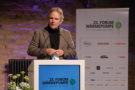Ein Mann spricht an einem Podium während des 23. Forums Wärmepumpe in Berlin. Er trägt einen grauen Mantel und hält einen Mikrofon. Im Hintergrund sind Sponsorenlogos sowie eine Pflanzenanordnung zu sehen.