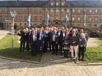 Eine Gruppe von etwa 40 Personen posiert im Freien vor einem historischen Gebäude. Alle tragen formelle Kleidung. Einige Personen lächeln, während andere freundlich schauen. Im Hintergrund sind Fahnen und eine Treppe sichtbar. Die Szene vermittelt eine Atmosphäre der Zusammenarbeit und des Austauschs.