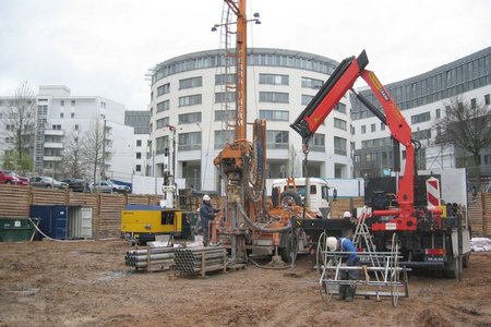 Bauplatz mit mehreren Maschinen, darunter ein Bohrgerät und ein Mobilkran. Arbeiter sind damit beschäftigt, Rohre und Materialien zu bewegen. Im Hintergrund sind moderne Gebäude sichtbar. Der Boden ist schlammig und uneben, was auf Bauarbeiten hinweist.