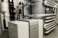 Das Bild zeigt ein Heizsystem mit einem großen weißen Gerät auf der linken Seite. Dahinter verlaufen zahlreiche Rohre, einige sind schwarz und orange markiert. Druckmesser sind an der Wand montiert, was auf ein technisches Umfeld hinweist.