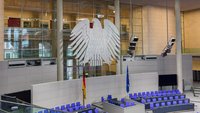 Der Plenarsaal des Deutschen Bundestags ist zu sehen, mit einem großen, weißen Bundesadler über der Sitzung. Es gibt blaue Stühle und Tische, sowie zwei Flaggen: die deutsche und die EU-Flagge. Der Raum strahlt eine moderne und offizielle Atmosphäre aus.