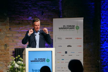 Ein Redner steht an einem Podium während des 23. Forums Wärmepumpe in Berlin. Er gestikuliert, während er spricht. Im Hintergrund ist eine Wand mit Sponsorenlogos und das Veranstaltungsschild sichtbar. Die Atmosphäre wirkt professionell und informativ.