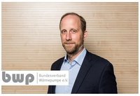 Ein Mann mit kurzen Haaren und einem Bart trägt einen dunklen Anzug und ein helles Hemd. Er steht vor einer neutralen, strukturierten Hintergrundwand. Im Vordergrund befindet sich das Logo des Bundesverbands Wärmepumpe e.V.