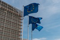Drei Flaggen der Europäischen Union wehen vor einem modernen Gebäudekomplex. Die Flaggen sind blau mit gelben Sternen und sind an Fahnenmasten befestigt, während der Himmel klar und sonnig ist.