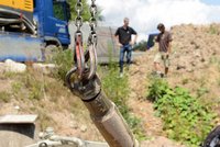 Das Bild zeigt eine nahaufgenommene Kranhakenaufhängung mit Ketten, die in einem Baustellenumfeld hängt. Im Hintergrund sind zwei Männer zu sehen, die auf einen Erdhügel blicken. Es vermittelt eine Arbeitsatmosphäre auf einer Baustelle.