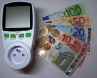 Ein Energieverbrauchsmesser liegt links im Bild. Daneben sind verschiedene Euro-Scheine und Münzen angeordnet. Die Scheine haben Werte von 5 bis 100 Euro. Münzen sind in unterschiedlichen Größen und Werten zu sehen.