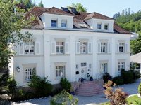 Eine elegante, zweistöckige Villa mit weißen Wänden und einem Satteldach. Die Fassade ist mit Fensterläden ausgestattet, und eine Schaufelstraße führt zu der Eingangstür. Vor dem Haus befinden sich gepflegte Gärten mit Bäumen und Sträuchern.