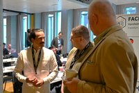 Drei Männer stehen in einem modernen Raum und führen ein Gespräch. Der Mann links trägt ein helles Hemd und hat einen Schnurrbart, während die anderen beiden Männer ein Gespräch führen und einer von ihnen eine Tasse hält. Im Hintergrund sind Tische und weitere Personen sichtbar.