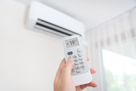 Eine Hand hält eine Fernbedienung für eine Klimaanlage, die auf eine Temperatur von 19 °C eingestellt ist. Im Hintergrund ist die Klimaanlage an der Wand sichtbar. Weiche Vorhänge lassen Licht herein.