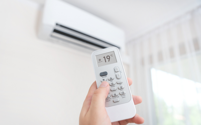 Eine Hand hält eine Fernbedienung für eine Klimaanlage, die auf eine Temperatur von 19 °C eingestellt ist. Im Hintergrund ist die Klimaanlage an der Wand sichtbar. Weiche Vorhänge lassen Licht herein.