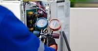 Eine Person in blauer Arbeitskleidung überprüft eine Klimaanlage. In der Hand hält sie ein Messgerät mit zwei Manometern in Blau und Braun, die den Druck anzeigen. Der Hintergrund zeigt die innere Elektronik der Klimaanlage.