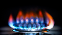 Ein naher Blick auf eine Gasflamme auf einem Herd. Die Flamme zeigt verschiedene Farben, hauptsächlich Blau und Orange, und hat eine spitze Form. Der Hintergrund ist dunkel, was den Kontrast zur Helligkeit der Flamme verstärkt.