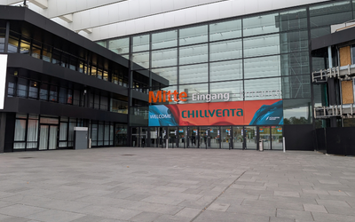 Eingangsbereich eines modernen Gebäudes mit großen Glasfenstern. Über dem Eingang befindet sich ein Banner mit der Aufschrift „Mitte Eingang“ und „WELCOME CHILLVENTA“. Im Vordergrund ist der Platz mit grauen Steinfliesen zu sehen.