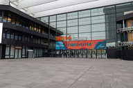 Eingangsbereich eines modernen Gebäudes mit großen Glasfenstern. Über dem Eingang befindet sich ein Banner mit der Aufschrift „Mitte Eingang“ und „WELCOME CHILLVENTA“. Im Vordergrund ist der Platz mit grauen Steinfliesen zu sehen.