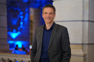 Ein Mann in einem grauen Anzug und blauen Hemd steht lächelnd an einer Säule. Im Hintergrund sind schwaches bläuliches Licht und weitere Personen sichtbar, die sich in einem eleganten Raum aufhalten.