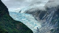 Ein majestätischer Gletscher erstreckt sich zwischen hohen, steilen Bergen. Die Eismasse leuchtet in blauen und weißen Tönen, während moosbedeckte Hänge den Gletscher umrahmen. Über dem Gletscher ziehen graue Wolken, die die beeindruckende Berglandschaft umgeben.