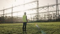 Eine Person in gelber Sicherheitsweste steht auf einer grünen Wiese und blickt auf eine stark strukturierte elektrische Umspannstation mit vielen Masten und Kabeln im Hintergrund. Die Sonne scheint, was eine helle Atmosphäre schafft.