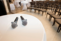 Zwei Mikrofone liegen auf einem runden Tisch in einem Veranstaltungsraum. Im Hintergrund sind Tische und Stühle für eine Versammlung zu erkennen. Die Atmosphäre wirkt bereit für eine Präsentation oder ein Event.