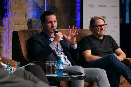 Zwei Männer sitzen auf einer Couch in einem Gespräch. Der Mann links, mit einem Mikrofon in der Hand, gestikuliert während er spricht. Der Mann rechts hört aufmerksam zu. Im Vordergrund sind Wasserflaschen und Gläser sichtbar. Die Umgebung hat eine moderne, lichtdurchflutete Atmosphäre.