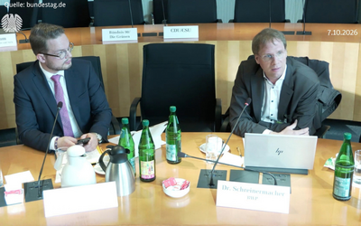Zwei Männer sitzen an einem Tisch in einem Sitzungssaal. Vor ihnen stehen Wasserflaschen und Snacks. Der Mann rechts spricht und hat einen Laptop vor sich. Der Sitzungstisch ist ordentlich und gut beleuchtet.