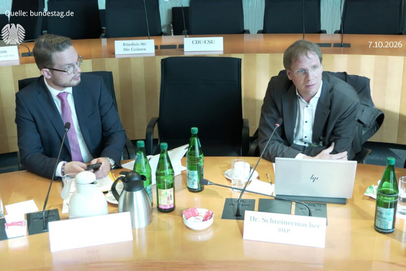 Zwei Männer sitzen an einem Tisch in einem Sitzungssaal. Vor ihnen stehen Wasserflaschen und Snacks. Der Mann rechts spricht und hat einen Laptop vor sich. Der Sitzungstisch ist ordentlich und gut beleuchtet.