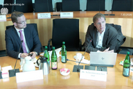Zwei Männer sitzen an einem Tisch in einem Sitzungssaal. Vor ihnen stehen Wasserflaschen und Snacks. Der Mann rechts spricht und hat einen Laptop vor sich. Der Sitzungstisch ist ordentlich und gut beleuchtet.