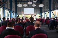 Ein Konferenzraum mit einer Gruppe von Menschen, die einem Vortrag folgen. Zwei Redner stehen vorne, während das Publikum auf Stühlen sitzt und auf eine Präsentation an der Wand blickt. Große Fenster bieten Tageslicht und einen Blick nach draußen.