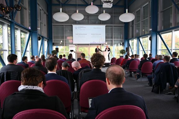 Ein Konferenzraum mit einer Gruppe von Menschen, die einem Vortrag folgen. Zwei Redner stehen vorne, während das Publikum auf Stühlen sitzt und auf eine Präsentation an der Wand blickt. Große Fenster bieten Tageslicht und einen Blick nach draußen.