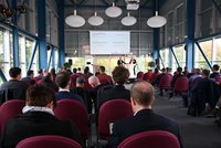 Ein Konferenzraum mit einer Gruppe von Menschen, die einem Vortrag folgen. Zwei Redner stehen vorne, während das Publikum auf Stühlen sitzt und auf eine Präsentation an der Wand blickt. Große Fenster bieten Tageslicht und einen Blick nach draußen.