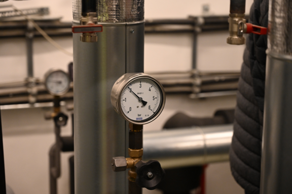 Manometer an einem Rohr, das den Druck anzeigt. Im Hintergrund sind weitere Rohre und ein Blick auf eine weitere Anzeige sichtbar. Die Umgebung deutet auf eine technische Einrichtung oder ein Versorgungssystem hin.