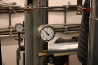 Manometer an einem Rohr, das den Druck anzeigt. Im Hintergrund sind weitere Rohre und ein Blick auf eine weitere Anzeige sichtbar. Die Umgebung deutet auf eine technische Einrichtung oder ein Versorgungssystem hin.