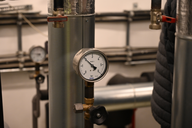 Manometer an einem Rohr, das den Druck anzeigt. Im Hintergrund sind weitere Rohre und ein Blick auf eine weitere Anzeige sichtbar. Die Umgebung deutet auf eine technische Einrichtung oder ein Versorgungssystem hin.
