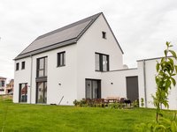 Das Bild zeigt ein modernes, weißes Einfamilienhaus mit einem Satteldach. Auf dem Dach sind Solarpanels installiert. Vor dem Haus befindet sich ein gepflegter Garten mit einer Sitzecke aus Holz und einem Grill, umgeben von grünem Rasen. Der Himmel ist bewölkt.