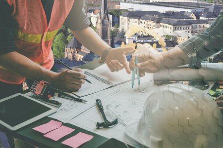 Zwei Personen in Arbeitskleidung stehen an einem Tisch mit technischen Zeichnungen und Bauplänen. Eine Person hält einen Bleistift, während die andere ein Maßband über die Pläne legt. Im Hintergrund sind städtische Gebäude und eine Kirche sichtbar.