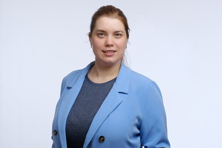 Eine Frau mit langen, blonden Haaren trägt eine blaue Jacke über einem grauen Oberteil. Sie steht vor einem einfarbigen, hellen Hintergrund und lächelt in die Kamera.