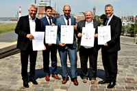 Fünf Männer stehen auf einer Steinstraße und halten Dokumente in den Händen. Sie tragen formelle Kleidung und lächeln in die Kamera. Im Hintergrund sind Gebäude und der Hafen sichtbar, was auf eine maritime Umgebung hinweist.