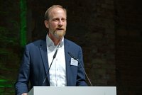 Ein Mann mit Vollbart hält eine Rede am Podium. Er trägt einen blauen Blazer und ein weißes Hemd. Die Kulisse ist eine Backsteinwand mit sanfter Beleuchtung. Der Mann wirkt engagiert und spricht zu einem Publikum.