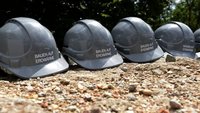 Sechsmal graue Schutzhelme liegen auf dem Boden. Auf jedem Helm steht der Schriftzug „BAUEN AUF ERDWÄRME“. Im Hintergrund sind Bäume zu erkennen. Die Szene vermittelt das Thema nachhaltiges Bauen mit Erdwärme.