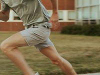 Ein sportlicher Mann in einem grauen T-Shirt und kurzen Hosen läuft aktiv über einen Rasen. Seine Beine sind in Bewegung, und er scheint sich auf ein Training oder einen Lauf im Freien zu konzentrieren. Der Hintergrund zeigt verschwommene Grasfläche und einige Gebäude.