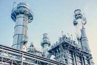 Die Abbildung zeigt mehrere große Industriekamine mit Metallkonstruktionen, die Dampf ausstoßen. Der Himmel im Hintergrund ist klar und blau. Die Anlagen wirken modern und industriell, mit Leitungen und Plattformen, die zu den Kaminen führen.