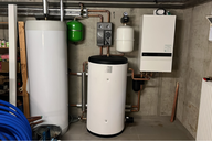 Abbildung eines Heizungsraums mit einer weißen Heizungsanlage, einem Wassertank, einem grünen Expansionsgefäß und verschiedenen Rohren. An der Wand befinden sich auch ein kleiner Wasserbehälter und ein Heizungssteuergerät. Im Vordergrund sind blaue Rohre zu sehen.